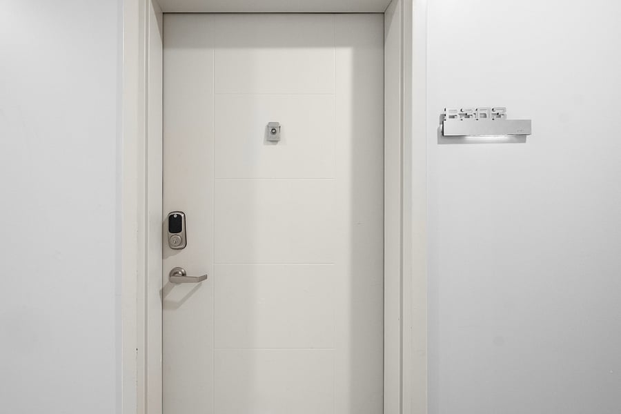 Sleek, modern white door set in a minimalist white wall. The door is equipped with a digital keypad lock and a peephole, emphasizing security and convenience. 