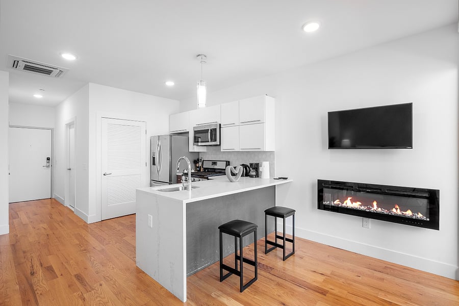 Stylish, open-concept kitchen and living area with white cabinetry, stainless steel appliances, and a sleek island with two black bar stools. Warm wood flooring contrasts with the clean white walls, while a wall-mounted TV and modern gas fireplace add a cozy, contemporary touch.

