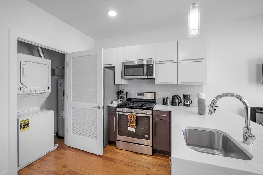 Compact, modern kitchen with white and dark cabinetry, stainless steel appliances, and a sleek marble countertop. To the left, an open closet reveals a stacked washer and dryer, blending laundry and kitchen functions in a clean, efficient layout. Light wood flooring and minimalist design enhance the bright, contemporary feel