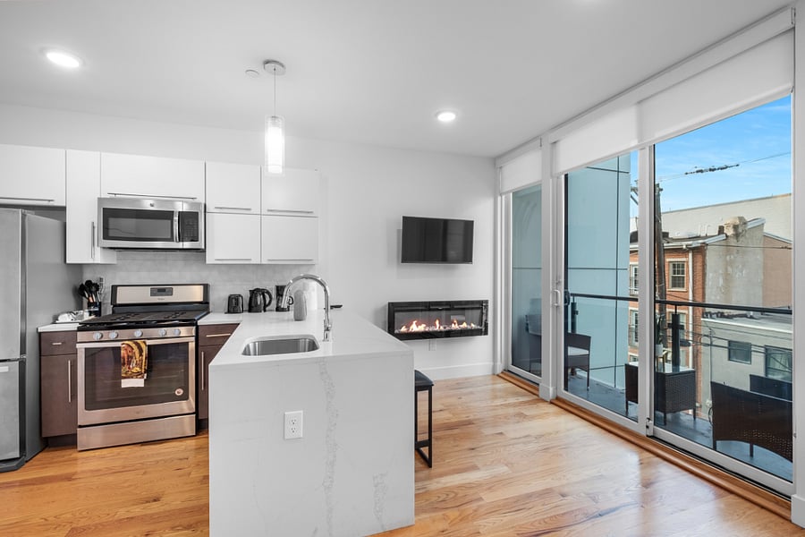 Bright, modern kitchen and living area with sleek stainless steel appliances, a stylish island with a marble countertop, and large windows that flood the space with natural light. The clean design and warm hardwood floors create a cozy yet contemporary vibe.