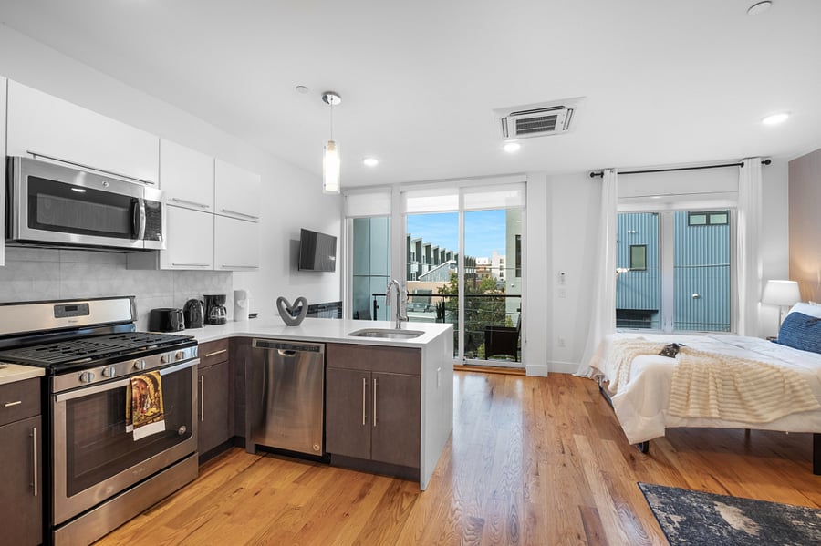 Bright, modern studio apartment with an open layout that combines kitchen, living, and bedroom areas. It features sleek cabinetry, stainless steel appliances, a central island with a sink, and warm hardwood floors. Large windows and a glass door lead to a balcony, filling the space with natural light and offering a view of nearby buildings.