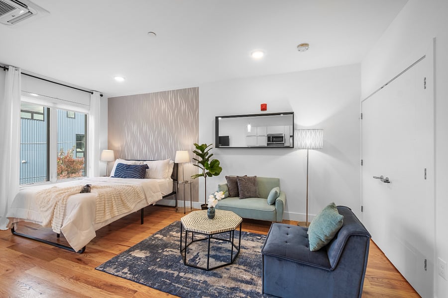Cozy, modern studio apartment with a stylish mix of textures and colors. It features a neatly made bed near a large window, a patterned accent wall, and a seating area with a green loveseat and a navy blue chair around a hexagonal coffee table. Warm wood flooring and soft lighting create a welcoming, contemporary atmosphere.