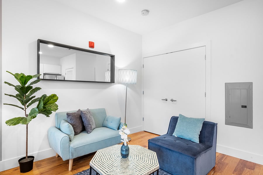 Cozy, modern living room with a minimalist design. It features a light blue loveseat and a dark blue accent chair, both adorned with plush pillows. A geometric-patterned coffee table sits between them, topped with a small floral arrangement. A tall floor lamp and a large wall mirror add depth and light to the space, while a potted plant brings in a touch of greenery. The room is bright and inviting, with white walls and warm wood flooring.
