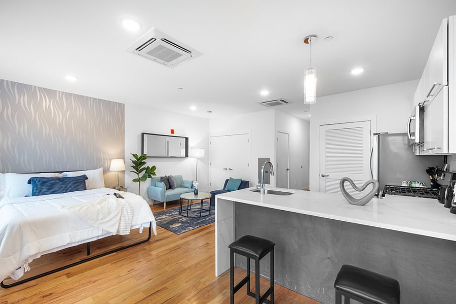 Bright, modern studio apartment with an open layout that combines kitchen, living, and bedroom areas. It features sleek cabinetry, stainless steel appliances, a central island with a sink, and warm hardwood floors. Large windows and a glass door lead to a balcony, filling the space with natural light and offering a view of nearby buildings.