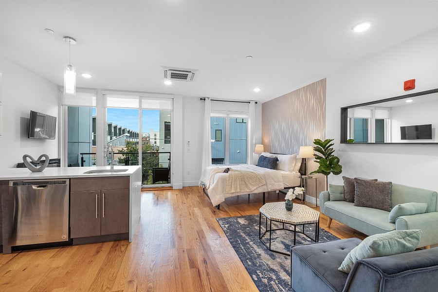 Bright, modern studio apartment with an open layout that combines kitchen, living, and bedroom areas. It features sleek cabinetry, stainless steel appliances, a central island with a sink, and warm hardwood floors. Large windows and a glass door lead to a balcony, filling the space with natural light and offering a view of nearby buildings.