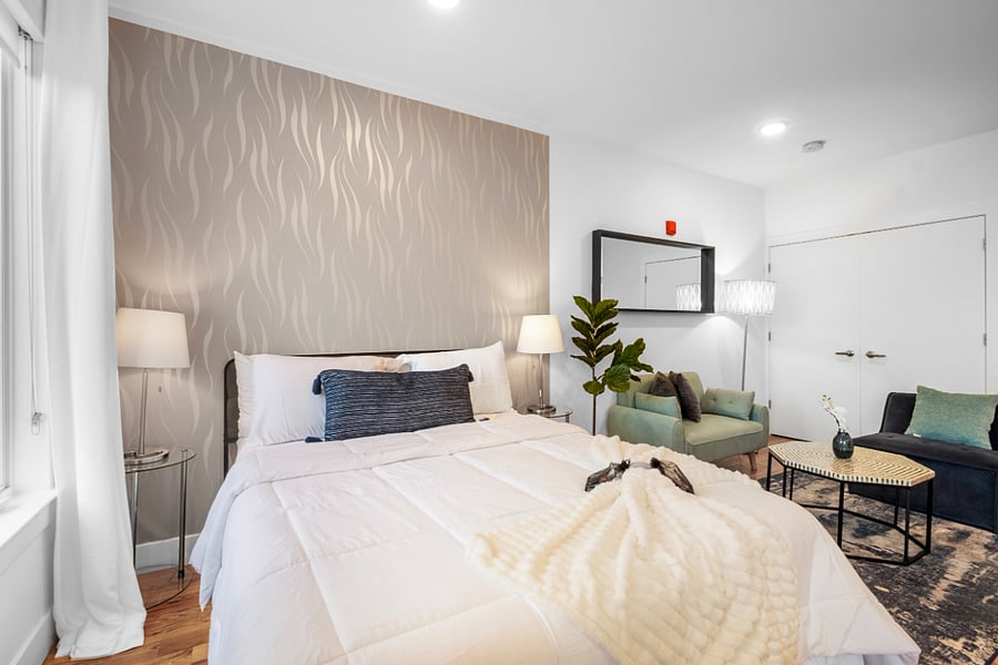 Bright, modern studio apartment with an open layout that combines kitchen, living, and bedroom areas. It features sleek cabinetry, stainless steel appliances, a central island with a sink, and warm hardwood floors. Large windows and a glass door lead to a balcony, filling the space with natural light and offering a view of nearby buildings.