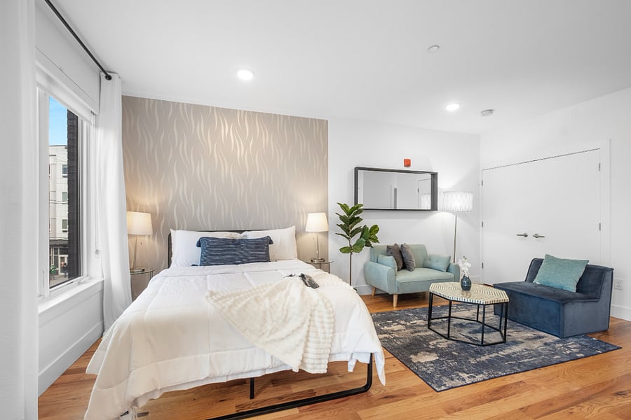 Bright, modern studio apartment with an open layout that combines kitchen, living, and bedroom areas. It features sleek cabinetry, stainless steel appliances, a central island with a sink, and warm hardwood floors. Large windows and a glass door lead to a balcony, filling the space with natural light and offering a view of nearby buildings.