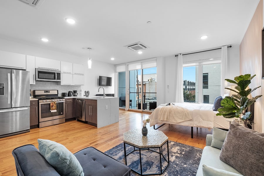Bright, modern studio apartment with an open layout that combines kitchen, living, and bedroom areas. It features sleek cabinetry, stainless steel appliances, a central island with a sink, and warm hardwood floors. Large windows and a glass door lead to a balcony, filling the space with natural light and offering a view of nearby buildings.