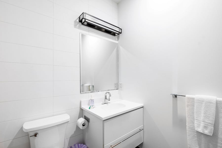 Sleek, modern bathroom with a minimalist aesthetic. It features a bathtub with a glass panel and wood-patterned tile accent wall, complemented by matching floor tiles. A white toilet and vanity with a built-in sink sit beneath a large mirror and a black light fixture. The clean white walls and subtle touches, like the purple waste bin, create a fresh and contemporary feel