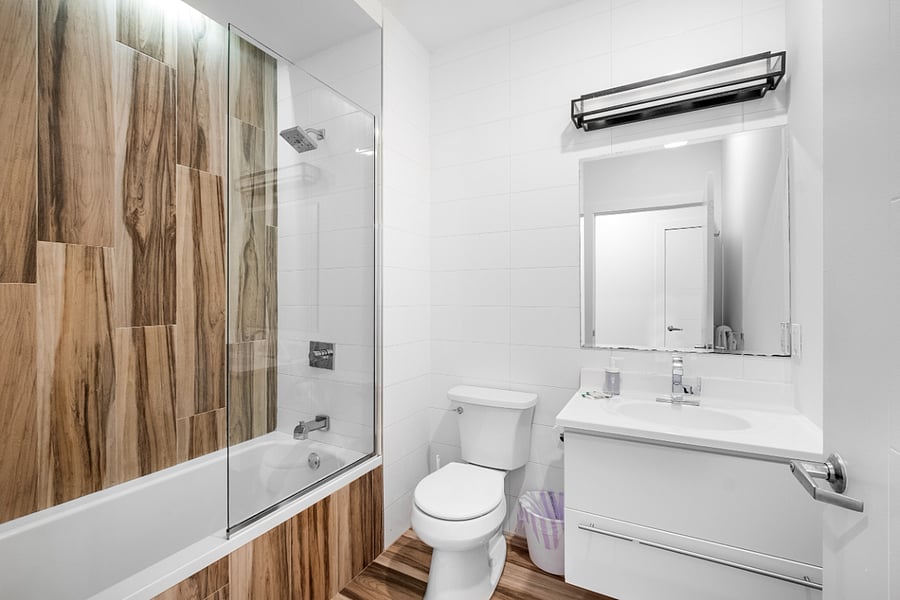 Sleek, modern bathroom with a minimalist aesthetic. It features a bathtub with a glass panel and wood-patterned tile accent wall, complemented by matching floor tiles. A white toilet and vanity with a built-in sink sit beneath a large mirror and a black light fixture. The clean white walls and subtle touches, like the purple waste bin, create a fresh and contemporary feel
