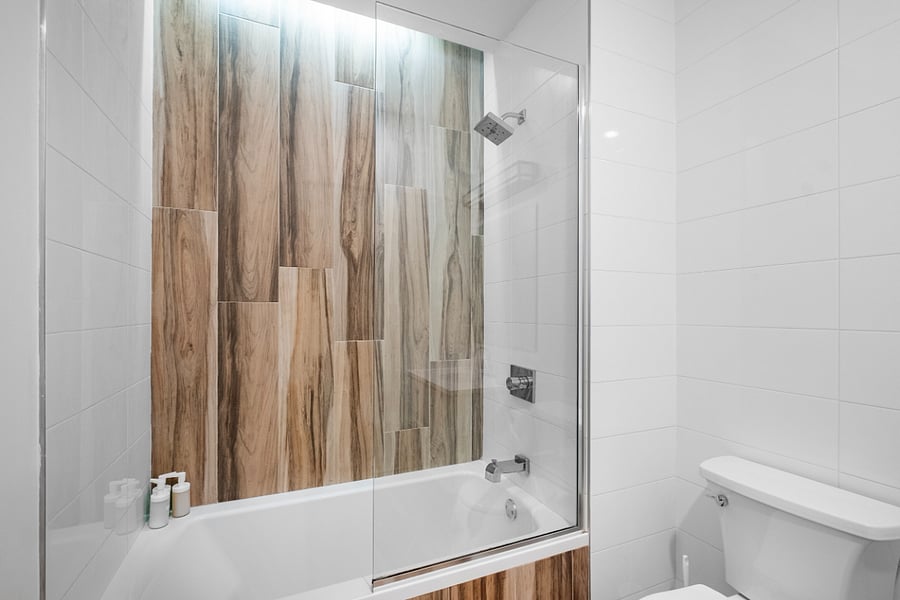Sleek, modern bathroom with a minimalist aesthetic. It features a bathtub with a glass panel and wood-patterned tile accent wall, complemented by matching floor tiles. A white toilet and vanity with a built-in sink sit beneath a large mirror and a black light fixture. The clean white walls and subtle touches, like the purple waste bin, create a fresh and contemporary feel