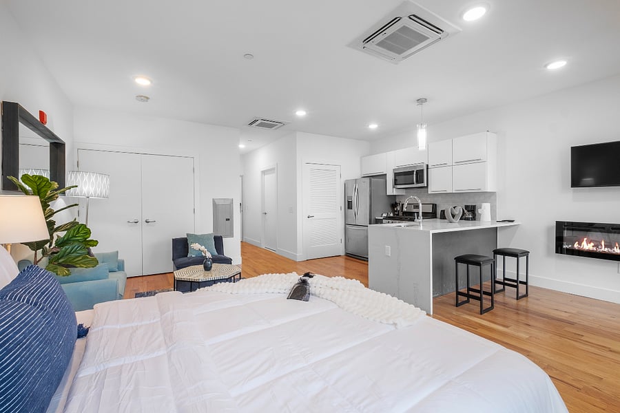 Stylish studio apartment with an open-concept layout. A neatly made bed with crisp white linens and blue accent pillows anchors the space, while a cozy sitting area with blue armchairs and a woven ottoman adds charm. The modern kitchen features stainless steel appliances, white cabinetry, and a sleek gray countertop with two black bar stools. A wall-mounted TV and electric fireplace complete the contemporary look, all set against light wood flooring and clean white walls.