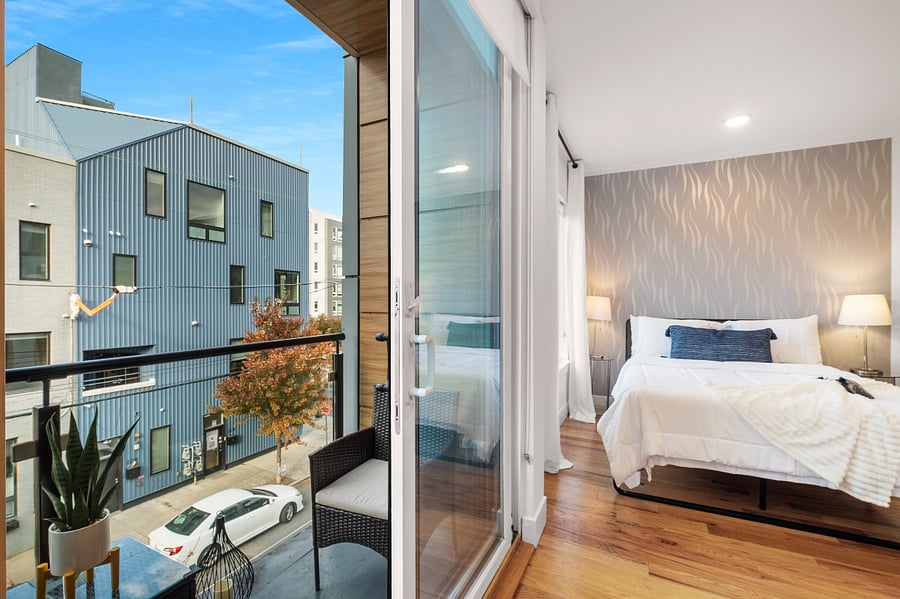 Bright, modern studio apartment with an open layout that combines kitchen, living, and bedroom areas. It features sleek cabinetry, stainless steel appliances, a central island with a sink, and warm hardwood floors. Large windows and a glass door lead to a balcony, filling the space with natural light and offering a view of nearby buildings.