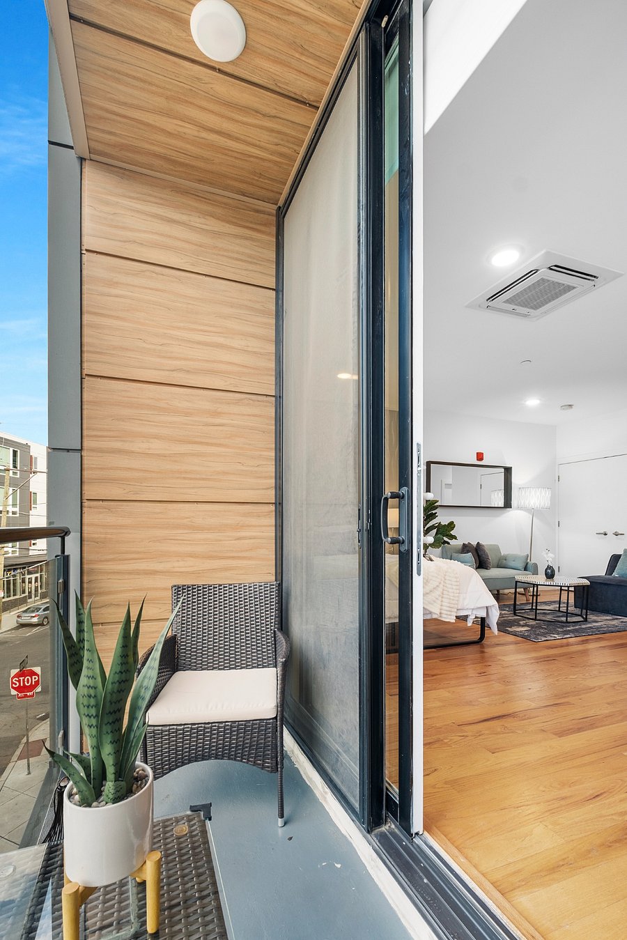 Stylish apartment balcony with wood-paneled walls and a sleek gray floor. A wicker chair with a white cushion and a small table holding a potted snake plant create a cozy outdoor nook. Through the open sliding glass door, a modern living room is visible, showcasing warm wood flooring, a white and dark blue couch, and recessed lighting.