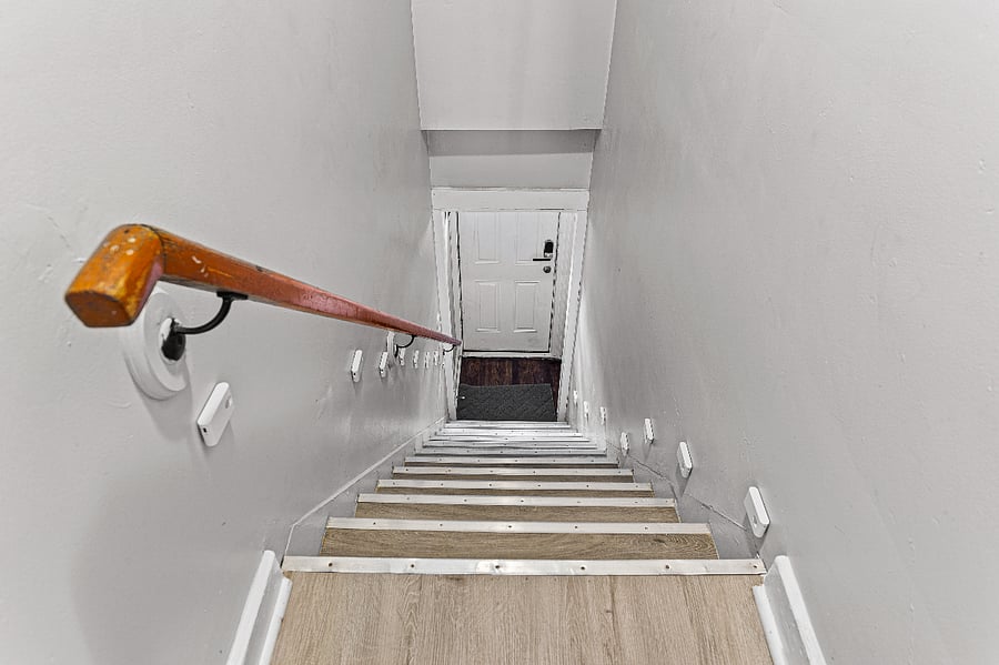 Apt. B Upper Flat on the other side of the white door looking down. Motion sensor lights & regular lighting illuminate the stairs.