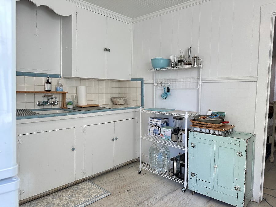 Rolling prep space. Coffee/tea light blue trolley. Cabinets have fixed storage shelves, and slightly askew older cabinet doors, a large selection of dishware, tupperware, cookware, and cooking basics.