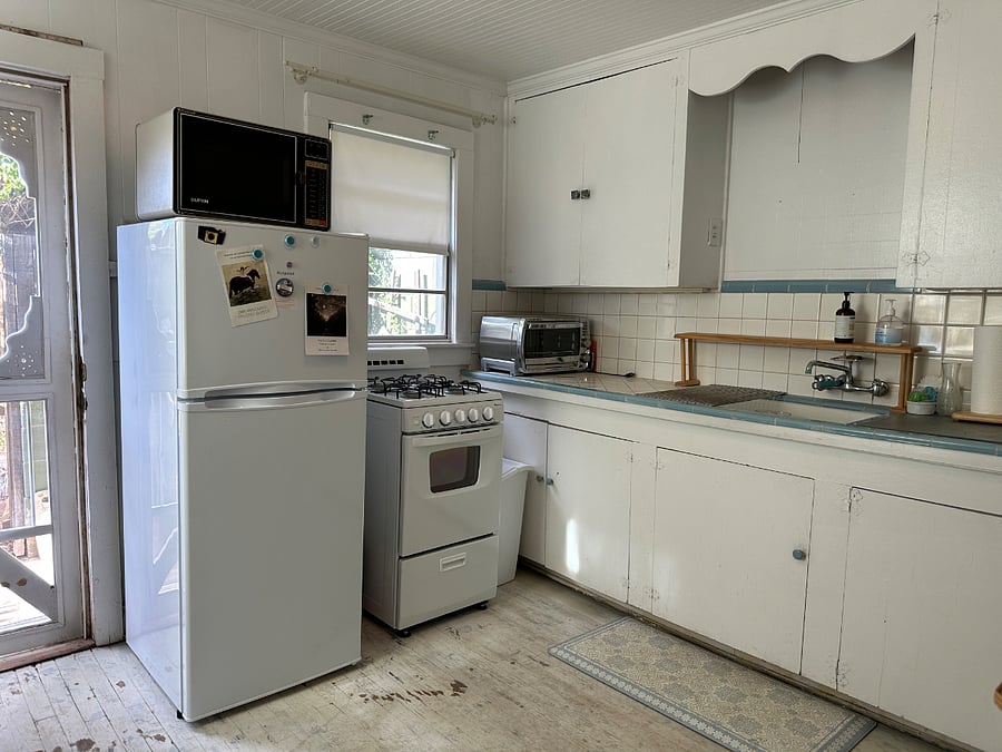 The full kitchen includes a 9 cu ft fridge, microwave, a 20-inch gas stove top with oven, the original large porcelain sink. Beautiful afternoon light. Handmade screen door to the tiny garden-fenced-in space.
