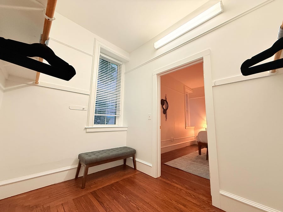 Spacious walk-in closet adjacent to the bedroom, featuring clothing racks, hangers, a window for natural light, and a convenient tufted bench.