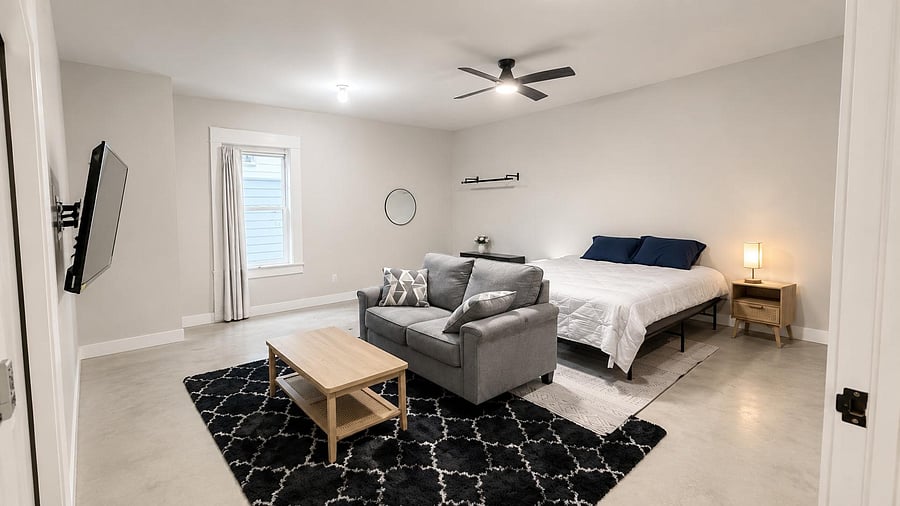 Spacious furnished bedroom with a king bed, seating area, wall-mounted TV, and ceiling fan, plus a window with curtains.