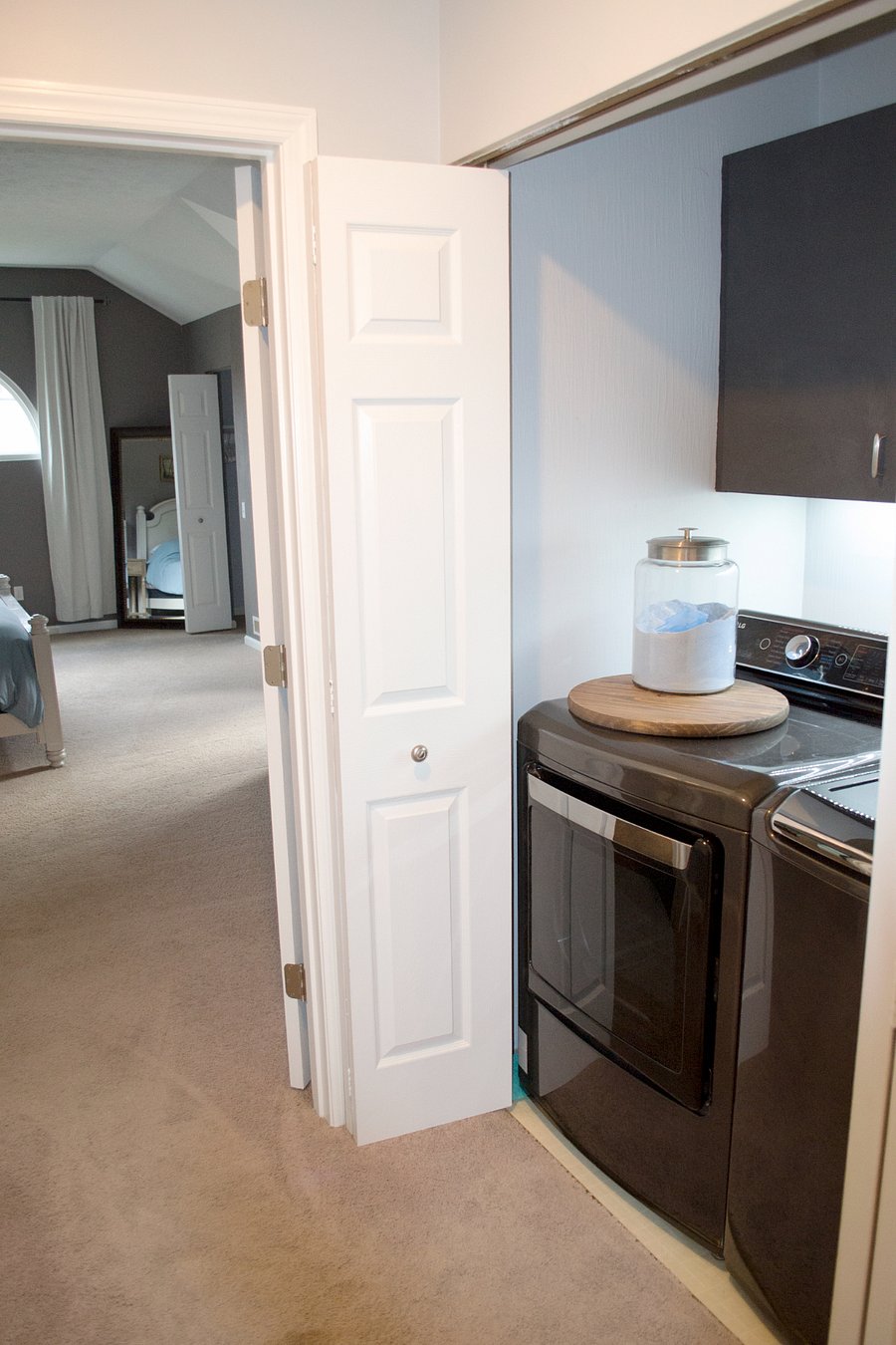 The laundry area in this home is on the second floor in the hallway next to the primary suite. Cleaning supplies are kept in the cabinets above the washer and dryer. There's also a smoke detector in this area as well. Directly across from the washer and dryer is a fold up table for folding clothes.