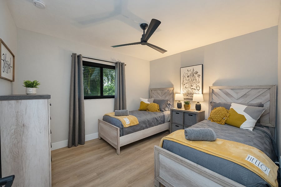 Two side-by-side twin beds with a dresser, lamps, ceiling fan, and window shades. Simple and comfy for kids or friends.