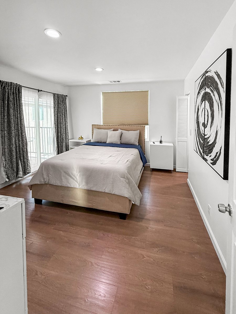 A clean, modern, hotel-calm master bedroom with soft neutrals, a plush upholstered bed, and a subtle blue accent. Bright natural light, minimal decor, and balanced symmetry give it a peaceful, polished feel.