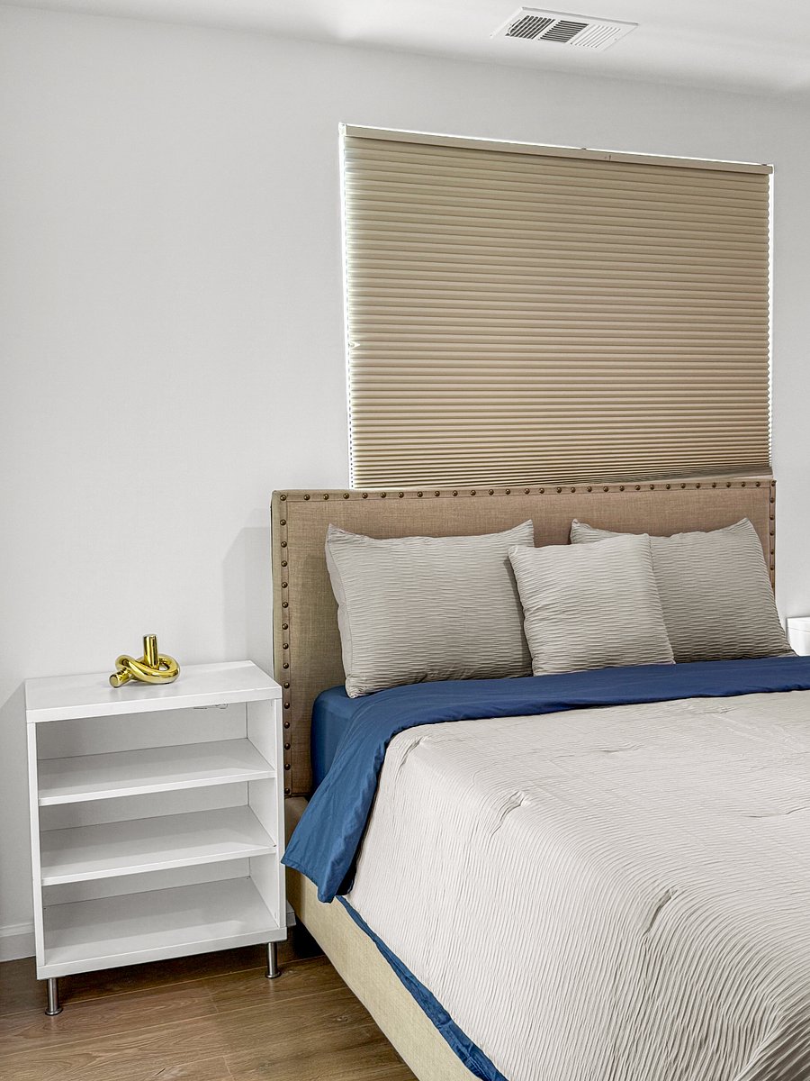 A clean, modern, hotel-calm master bedroom with soft neutrals, a plush upholstered bed, and a subtle blue accent. Bright natural light, minimal decor, and balanced symmetry give it a peaceful, polished feel.