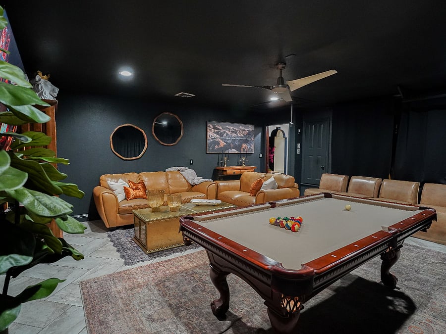 The Velvet Lounge: Moody Converted Garage Game Room Sanctuary
Step into the ultimate private escape a 400 sq ft converted garage transformed into an atmospheric velvet-lined lounge, where moody sophistication meets effortless entertainment in your curated ranch-style retreat.
Deep charcoal walls and dramatic blackout curtains create an intimate, illuminated by soft recessed lighting and a ceiling fan for perfect comfort. At the center, a professional 7.5 ft pool table commands the space, surrounded by plush Italian caramel leather sofa and loveseat ideal for sinking in with friends or unwinding solo. Partnered with brass side tables and vintage gold coffee tables.
Large layered rugs in rich patterns and textures bring the expansive room together, anchoring the seating areas with warmth, depth, and cohesive elegance while softening the space underfoot.

A massive 75" smart TV mounted on the wall delivers cinematic immersion, while a classic dartboard adds friendly competition. Tucked discreetly, a full-size fridge for your BYOB and chilled drinks ensures refreshments are always at hand.
Nestled thoughtfully within this indulgent haven, a laundry area is elegantly concealed behind coordinating curtains, blending seamless functionality with the room's refined aesthetic.
This velvet lounge is your personal retreat for memorable game nights, relaxed gatherings, or quiet evenings of pure restoration timeless ranch luxury redefined.
