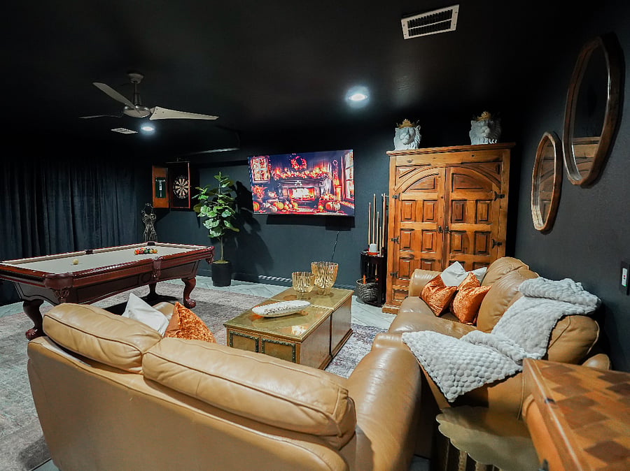The Velvet Lounge: Moody Converted Garage Game Room Sanctuary
Step into the ultimate private escape a 400 sq ft converted garage transformed into an atmospheric velvet-lined lounge, where moody sophistication meets effortless entertainment in your curated ranch-style retreat.
Deep charcoal walls and dramatic blackout curtains create an intimate, illuminated by soft recessed lighting and a ceiling fan for perfect comfort. At the center, a professional 7.5 ft pool table commands the space, surrounded by plush Italian caramel leather sofa and loveseat ideal for sinking in with friends or unwinding solo. Partnered with brass side tables and vintage gold coffee tables.
Large layered rugs in rich patterns and textures bring the expansive room together, anchoring the seating areas with warmth, depth, and cohesive elegance while softening the space underfoot.

A massive 75" smart TV mounted on the wall delivers cinematic immersion, while a classic dartboard adds friendly competition. Tucked discreetly, a full-size fridge for your BYOB and chilled drinks ensures refreshments are always at hand.
Nestled thoughtfully within this indulgent haven, a laundry area is elegantly concealed behind coordinating curtains, blending seamless functionality with the room's refined aesthetic.
This velvet lounge is your personal retreat for memorable game nights, relaxed gatherings, or quiet evenings of pure restoration timeless ranch luxury redefined.