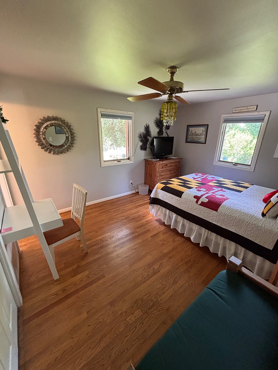 This photo shows the three drawer wooden dresser, the television, the two casement windows with pulldown screens and blackout shades, the queen size bed, and the storage bench and ceiling fan with light.