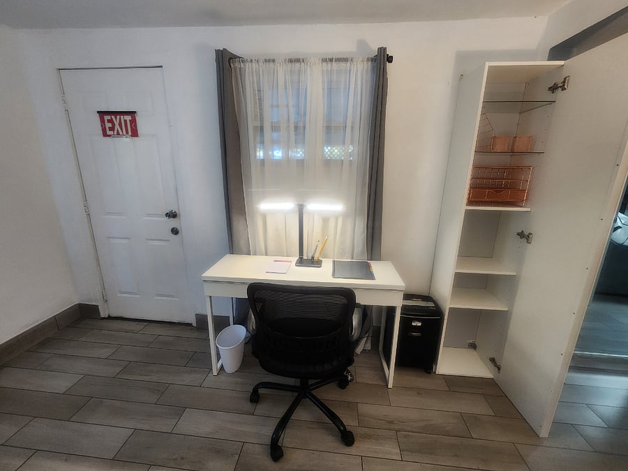 A refined workspace is arranged within the bedroom, offering a composed and efficient setting for productivity. The area includes a sleek storage cabinet, a modern desk with a focused task light, and a paper shredder to support privacy and professional needs. Just beside the office area, an exterior door provides direct access to the jacuzzi and the laundry area, creating a seamless flow between work, relaxation, and convenience.
