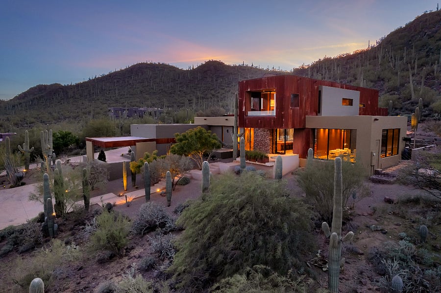 Twilight view of the home nestled into the hillside, with warm interior light glowing against the desert landscape.