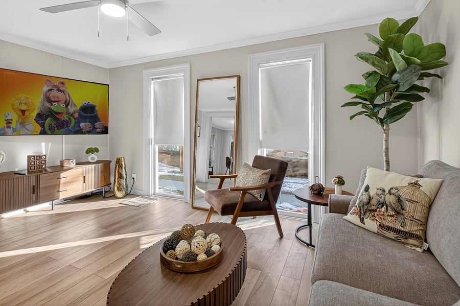 The living room features floor-to-ceiling windows that provide bright natural light, along with a sofa, two chairs, a lamp, a center table, a Smart TV, and a securely mounted leaning full-length mirror, creating a comfortable and inviting space.