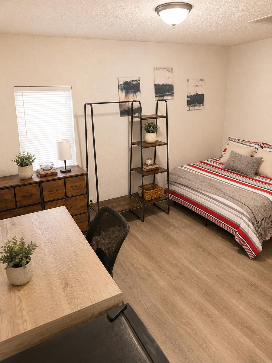 Furnished private bedroom with twin bed, desk workspace, dresser, and open shelving plus a window with blinds.