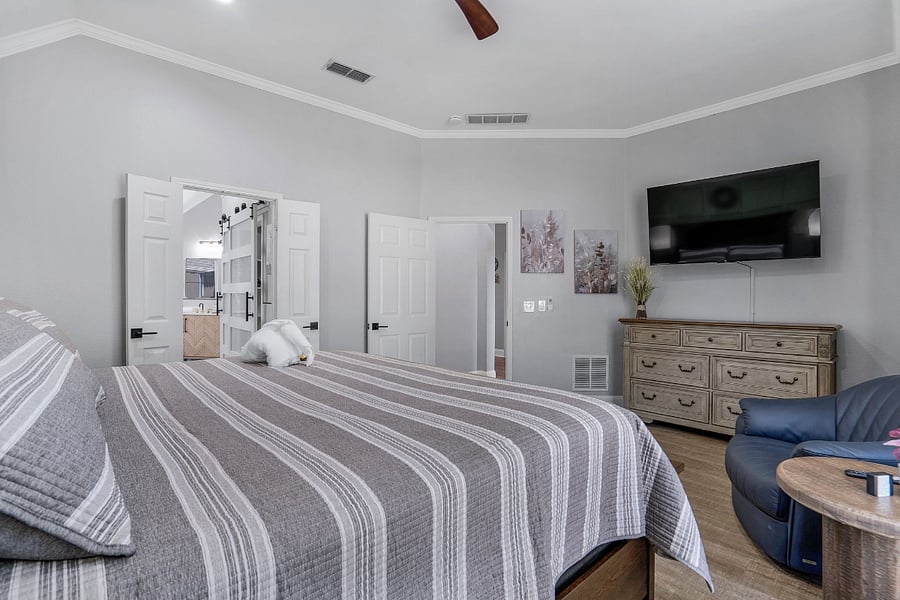 Primary bedroom has a king bed, with dresser, nightstands, lamps, ceiling fan, large smart TV, and on front of the house attached to primary walk in closet and bathroom