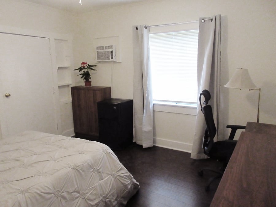 Private Bedroom, facing window from entrance door. Pictured are blackout curtains, built-in shelves, double-door bookshelf, mini-fridge, desk, ergonomic chair, and floor lamp.