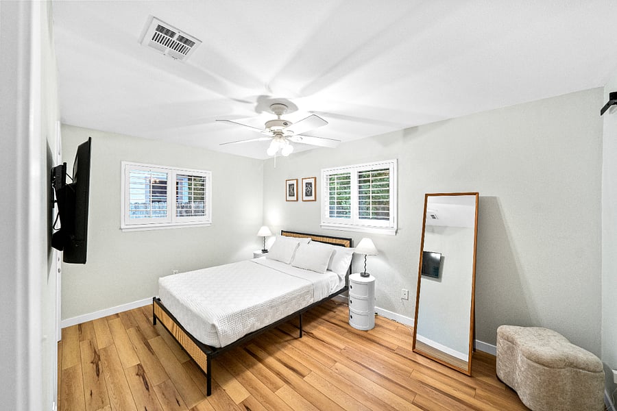 Primary bedroom wide angle shot of the room with the queen bed, full-length mirror, mounted smart TV, ottoman, dual nightstands and estate window shutters.