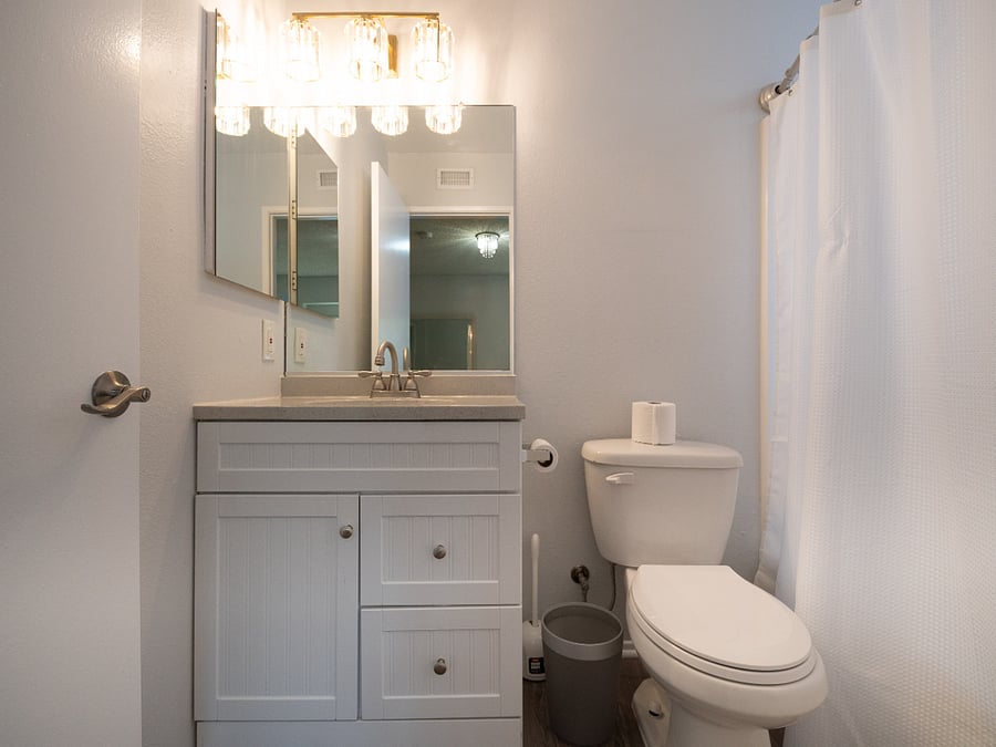 Bedroom 1 bathroom with shower curtain, garbage pail, toilet bowl brush, cleaning supplies.
