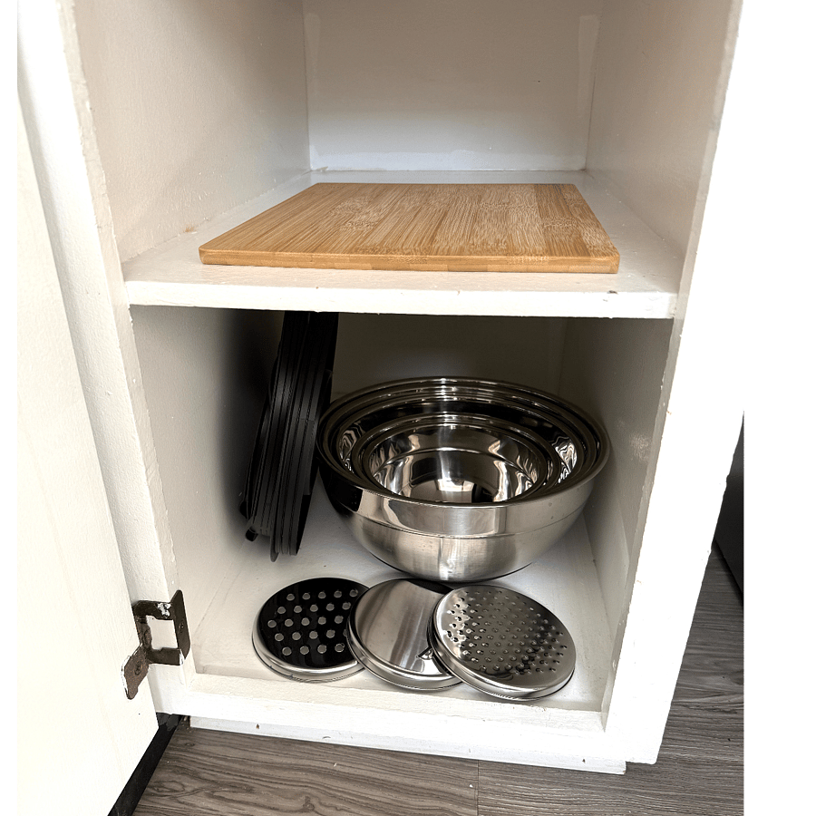 Five mixing bowls with lids and three different cheese graders/slicers that attach to one of the lids, bamboo cutting board in kitchen cabinet.