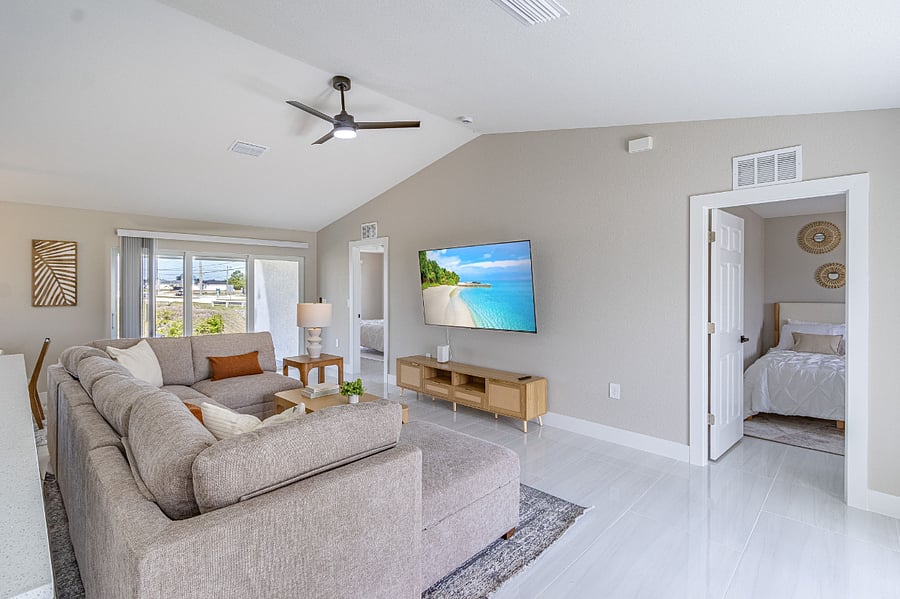 Bright and airy open-concept living room with vaulted ceilings, a large sectional sofa, wall-mounted smart TV, and French doors leading to the backyard. Fully furnished and move-in ready.