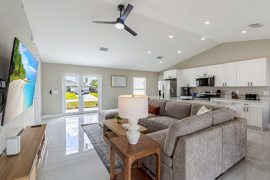 Wide-open great room showcasing the seamless flow from living area to kitchen perfect for entertaining. Features vaulted ceilings, a modern ceiling fan, large sectional, and direct access to the front of the house through glass French doors.