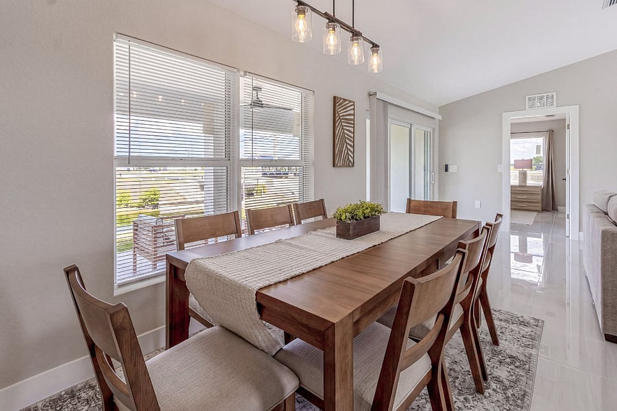 Elegant dining area with an 8-seat solid wood table, upholstered chairs, woven table runner, and a modern industrial pendant light. Natural light floods in through large windows overlooking the yard.