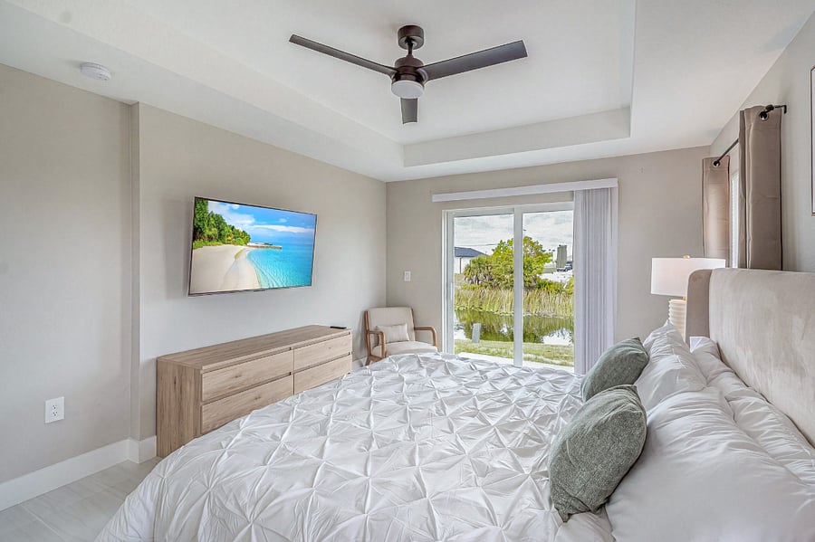 Primary suite with a king-size bed, wall-mounted smart TV, dresser, ceiling fan, and a private sliding glass door opening to the backyard with peaceful canal views. Serene and spacious.
