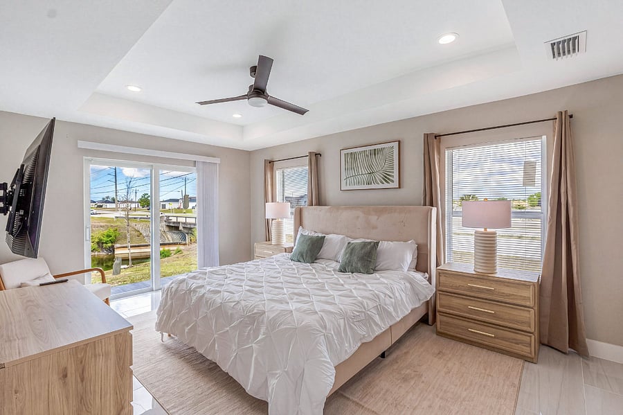 Primary suite with a king-size bed, wall-mounted smart TV, dresser, ceiling fan, and a private sliding glass door opening to the backyard with peaceful canal views. Serene and spacious.