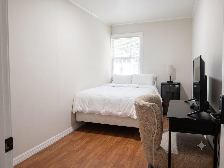 Furnished bedroom with a comfortable bed, side table and lamp, plus a dedicated desk setup and window with blinds.