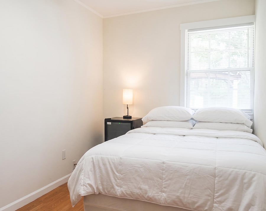 Furnished bedroom with a full-size bed, nightstand lighting, and a large window with blinds for comfortable daily living.