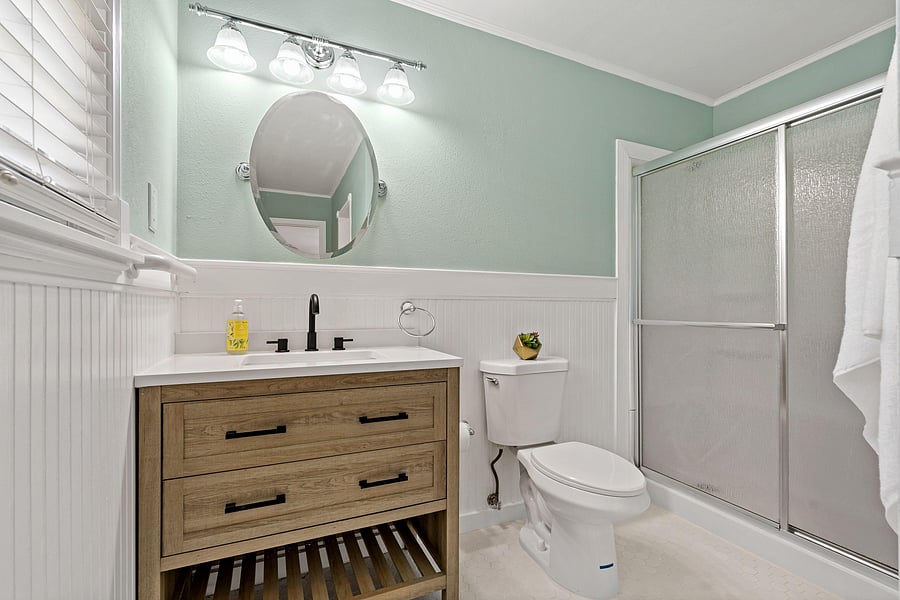Updated full bath with shaker-style vanity, solid-surface counter, and a glass-enclosed tub/shower with overhead lighting.