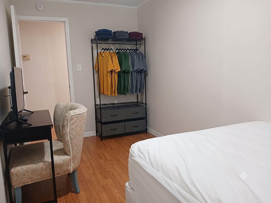 Furnished bedroom with wood-look flooring, a work desk and TV, and open clothing storage with drawer organizers.
