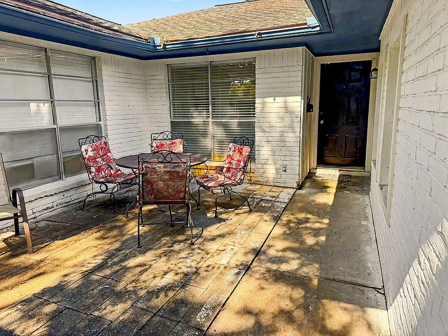 Covered patio entry with paved seating area, window shutters, and space for outdoor dining or relaxing near the front door.