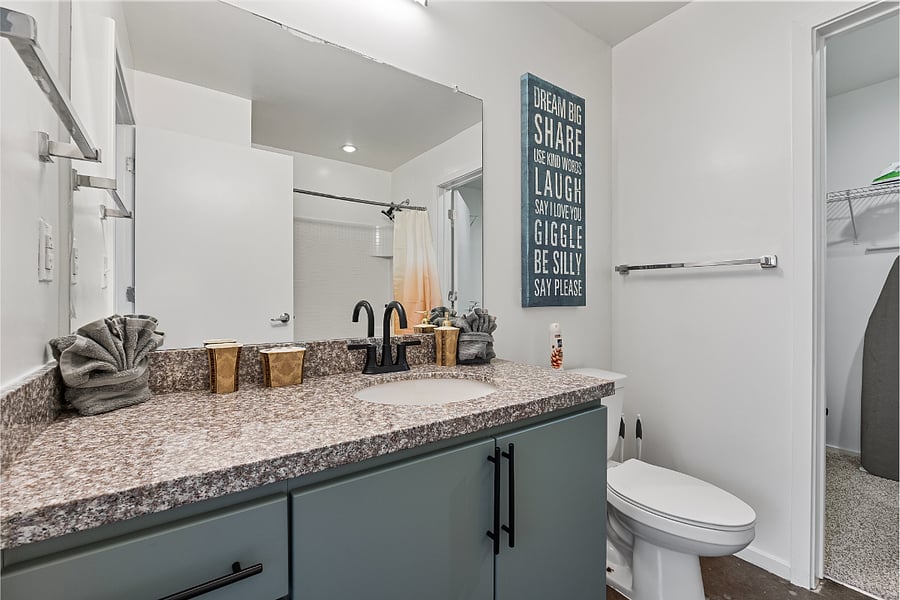 The modern bathroom features a large vanity with ample storage space, a spacious mirror, and a sleek commode. Designed with both style and functionality in mind, this clean and bright space provides plenty of room to prepare for your day.